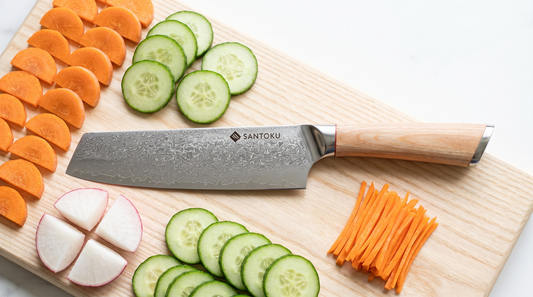 Overhead view of a Santoku Knives nakiri on a pale board surrounded by carrot half-moons, cucumber rounds, daikon quarters and fine matchsticks