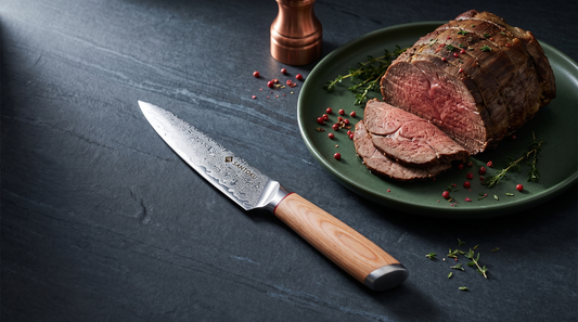 Haruta 5 inch VG10 Damascus utility knife with wooden scabbard on dark slate beside carved roast beef on a green stoneware platter